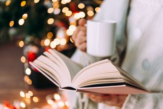 A person enjoying a book while drinking a coffee in front of a Christmas tree.