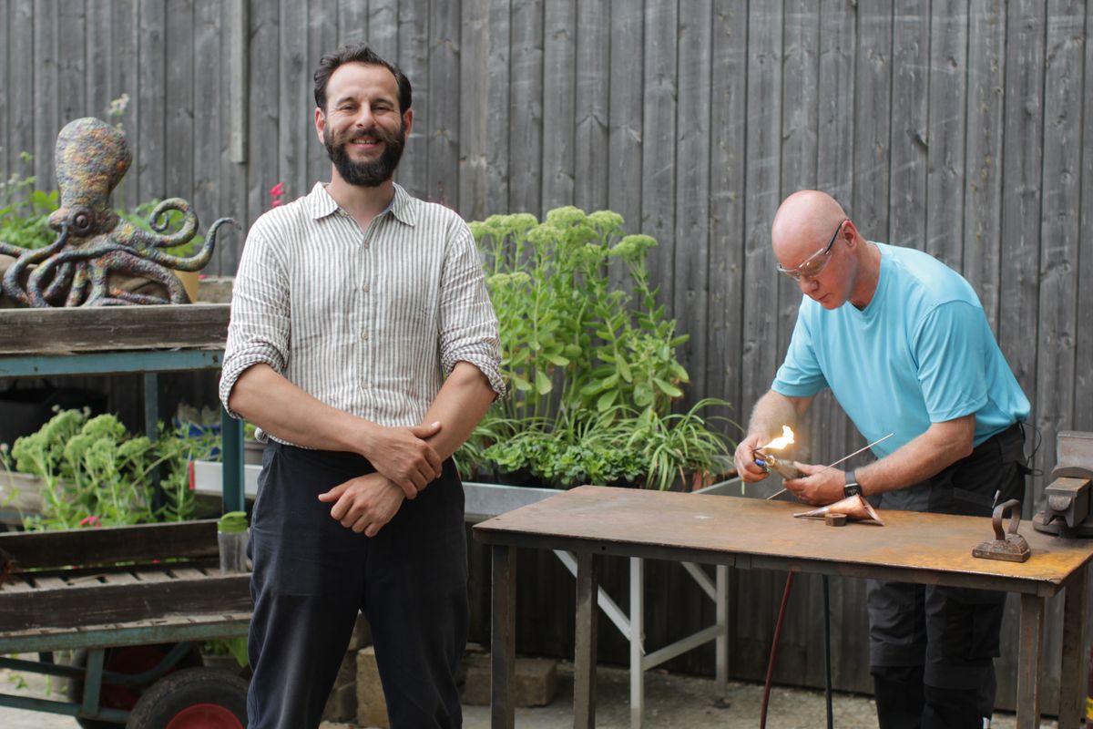 Dominic Chinea smiling as coppersmith Roy works behind him. 