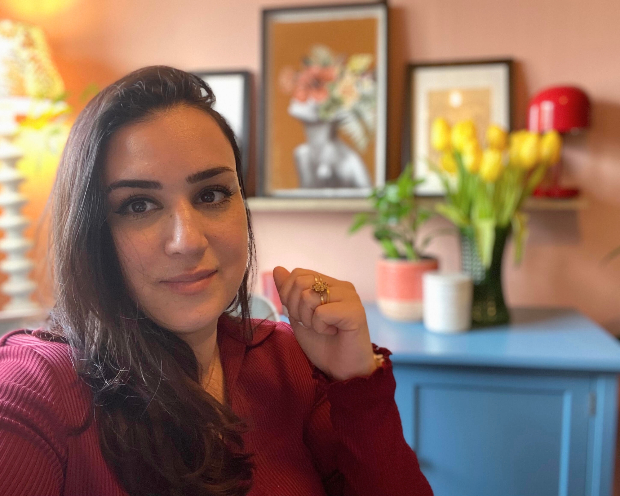 Punteha, a middle eastern woman with dark long hair, in a red long sleeve dress, in her small terracotta home office with lamp, blue sideboard and plants in background