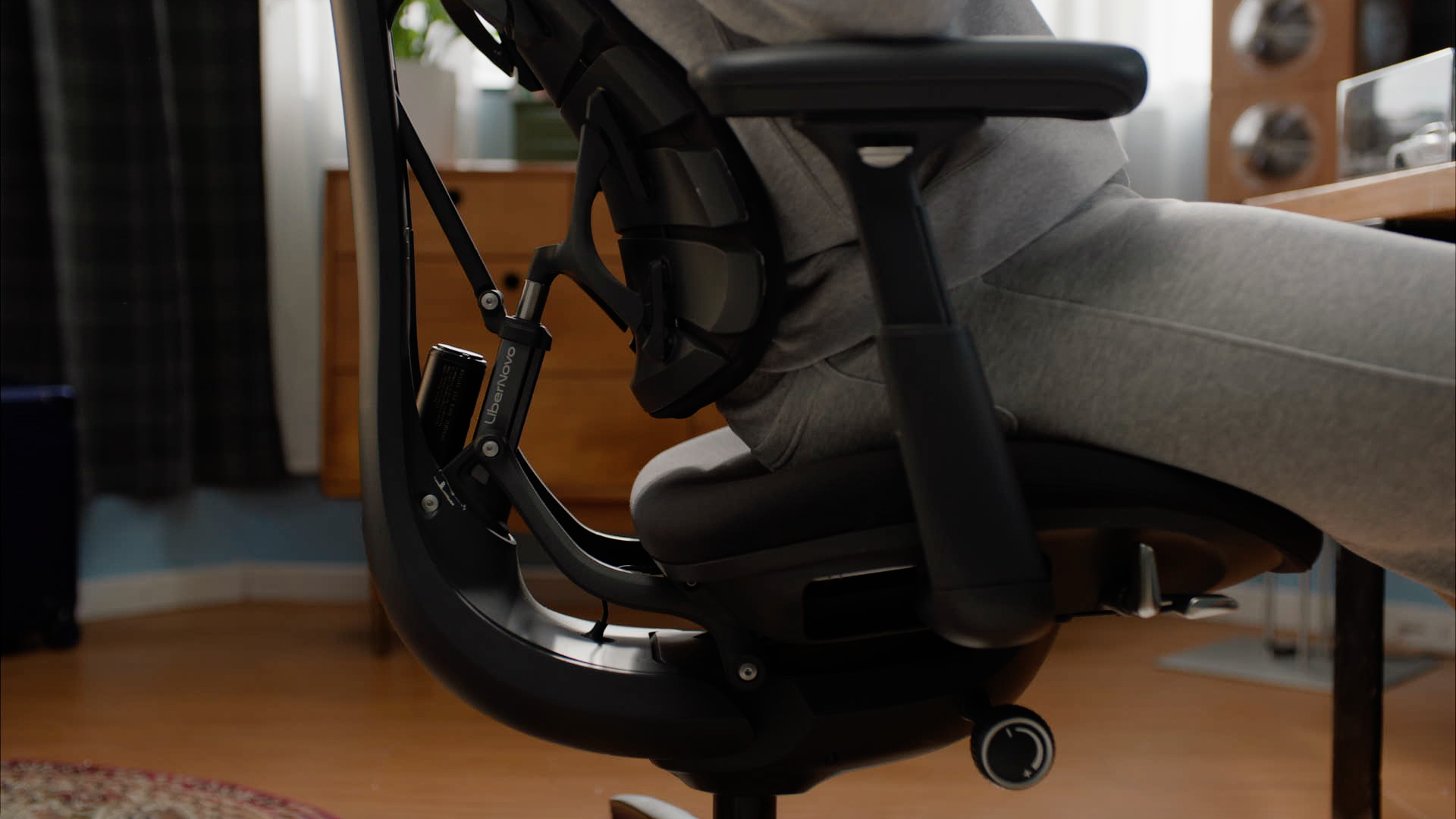 Close-up side view of a person reclining in a black ergonomic office chair, showing the chair&amp;rsquo;s back support, frame, and adjustment hardware in a home office setting.
