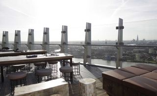 A rooftop sitting area with long wooden tables, round chairs, leather benches and a glass railing overlooking the city.
