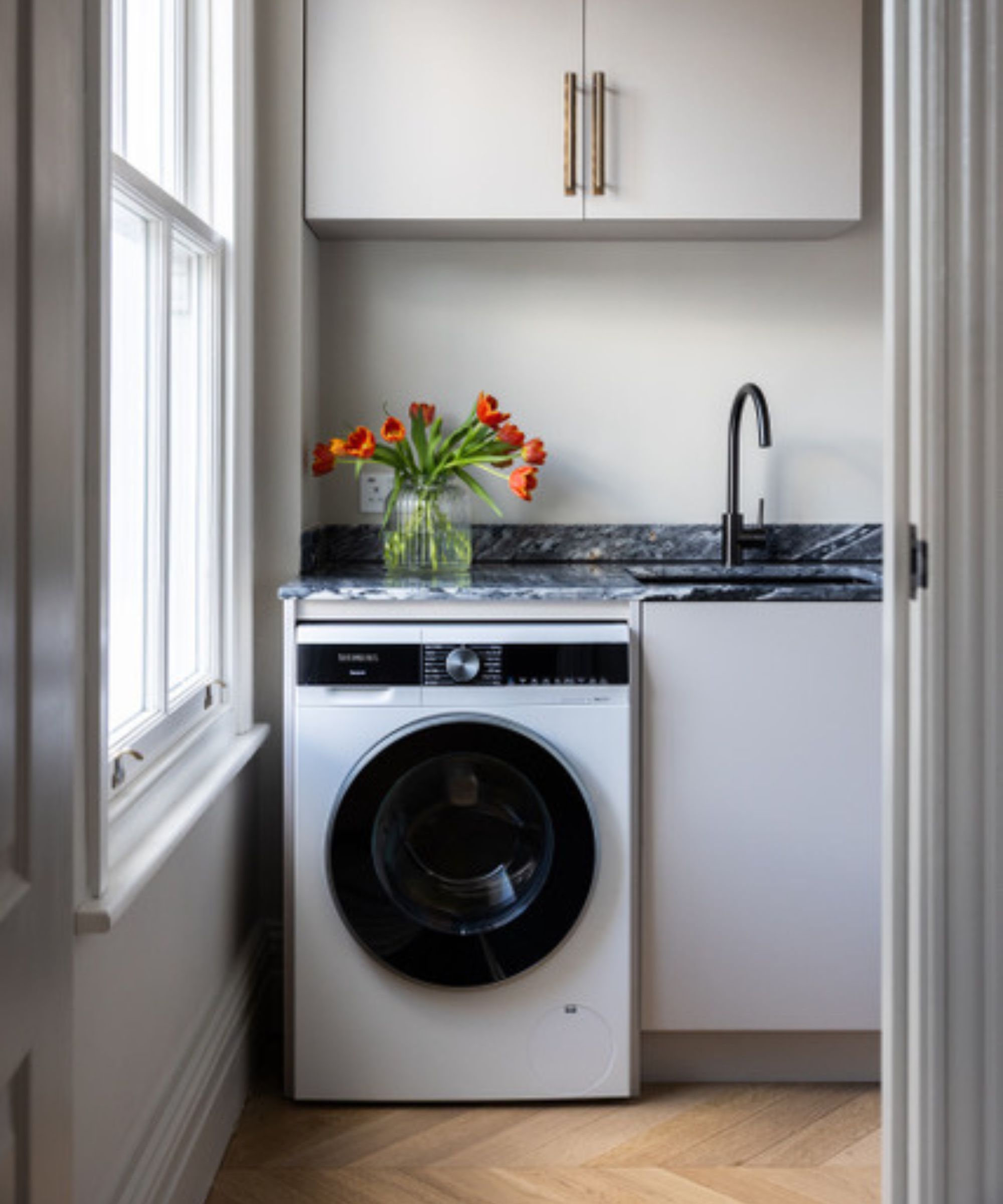A compact utility room with white cabinetry, a matching dark marble worktop and sink, black tap, washing machine and herringbone wood floor, with orange tulips adding a pop of colour.