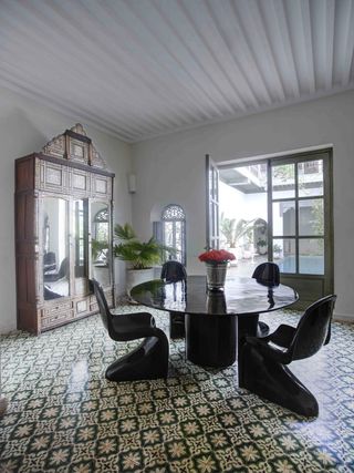 A hotel dining area with a round black table, black chairs, a large cupboard with mirrors on the doors, a potted plant, patterned floor tiles and large double doors to a courtyard.