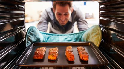 View from rear of oven of tray of salmon fillets and man in gray button-up shirt with hands in oven gloves.