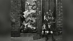 Black and white photograph of children playing on a rope swing wedged between two brick walls, with two more boys crouched nearby covering their faces, taken in
