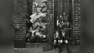 Black and white photograph of children playing on a rope swing wedged between two brick walls, with two more boys crouched nearby covering their faces, taken in Byker, Tyneside, in 1976.