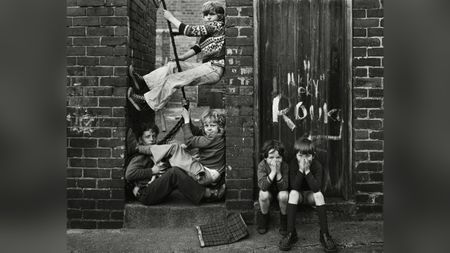 Black and white photograph of children playing on a rope swing wedged between two brick walls, with two more boys crouched nearby covering their faces, taken in Byker, Tyneside, in 1976.