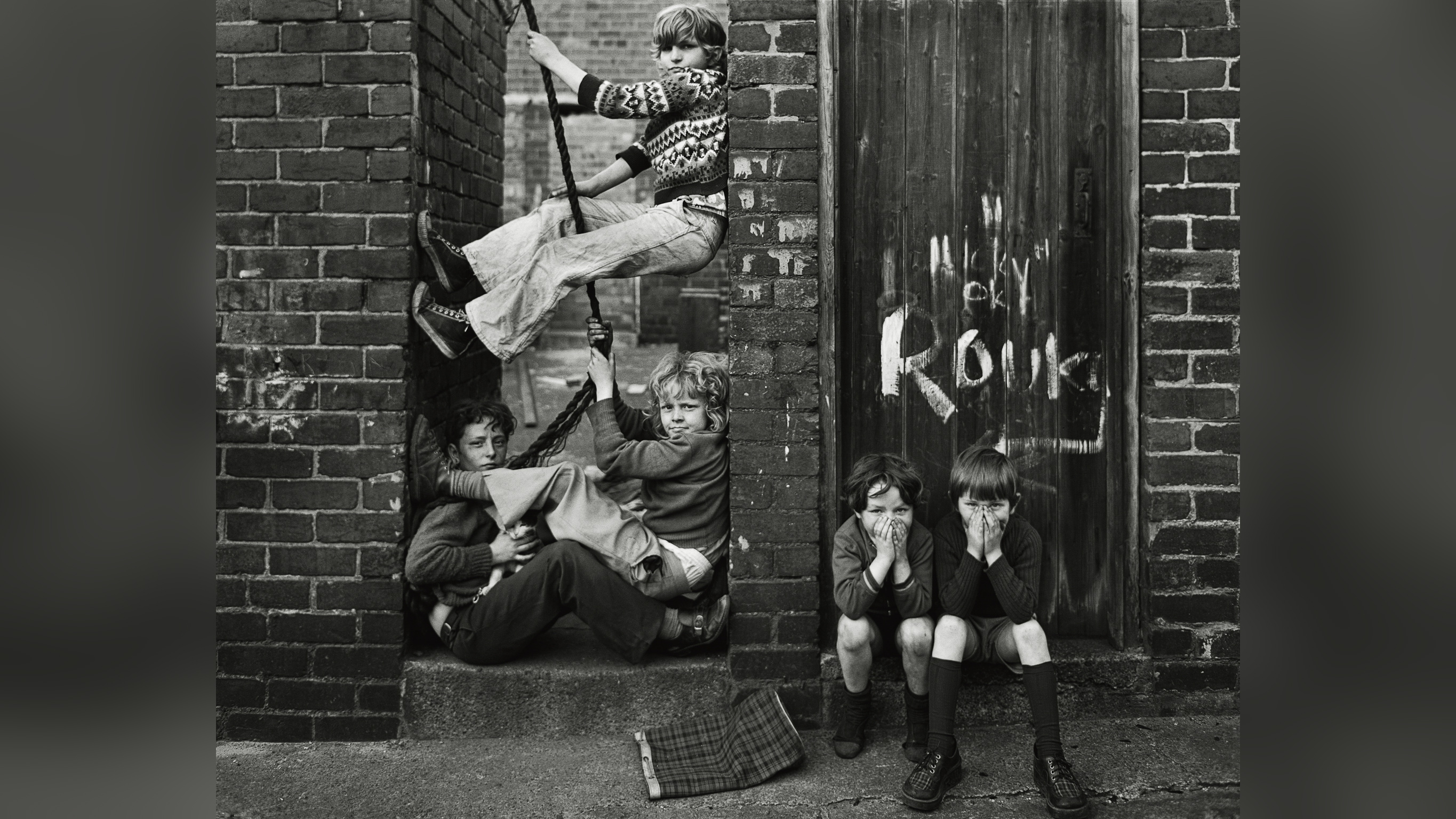 Black and white photograph of children playing on a rope swing wedged between two brick walls, with two more boys crouched nearby covering their faces, taken in Byker, Tyneside, in 1976.