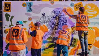 Four men in high visibility jackets assembling graffiti, art and bright stickers onto a large white board