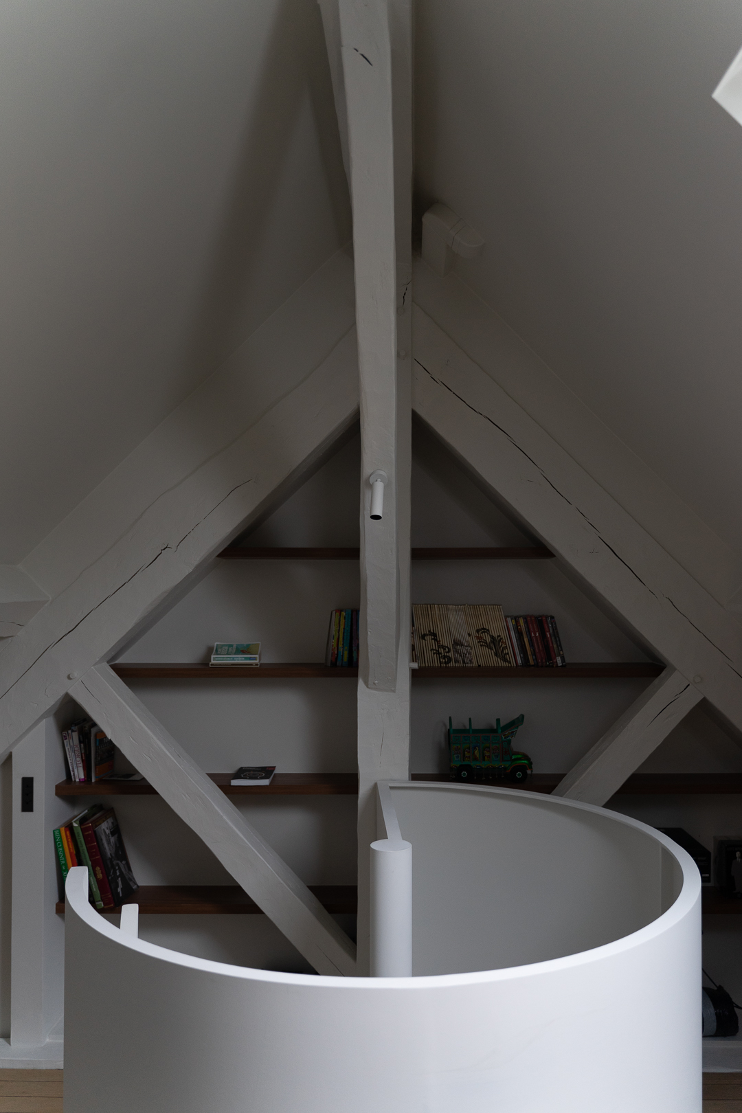 A refined attic apartment interior characterized by a warm, modernist aesthetic. The space is organized as an open-plan living area under a pitched roof, with exposed white-painted beams and triangular dormer windows that bring in soft natural light.