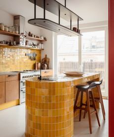 Yellow tile sinuous kitchen island with wooden stools and lighter yellow tiles backsplash under open shelving