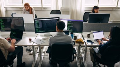 Male and female software developers working on desktop computers and laptops in an open plan office space with source code pictured on desktop monitor.