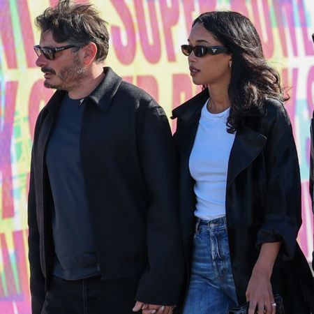 Laura Harrier walks outside wearing high-waisted straight-leg jeans with heeled boots, a white tee and a black trench coat.