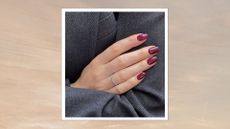 A close-up of someone wearing a blazer, with their arms crossed and their hand pictured with short burgundy almond nails by nail artist, Mateja Novakovic (@matejanova)/ in a beige to grey sunset-style template