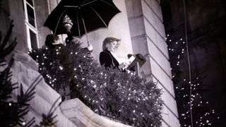 Princess diana stood on a balcony in front of a podium on Regents street in London with shrubery around the buidling