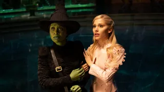 Cynthia Eviro as Elphaba and Ariana Grande as Glinda. They stand side by side and hold onto each other, looking off camera at something.