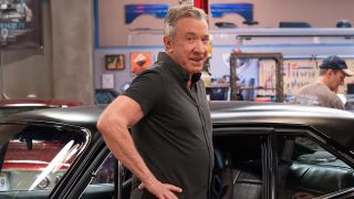 Tim Allen in a matte black shirt standing by a matte black hot rod on the set of the car shop in Shifting Gears. 