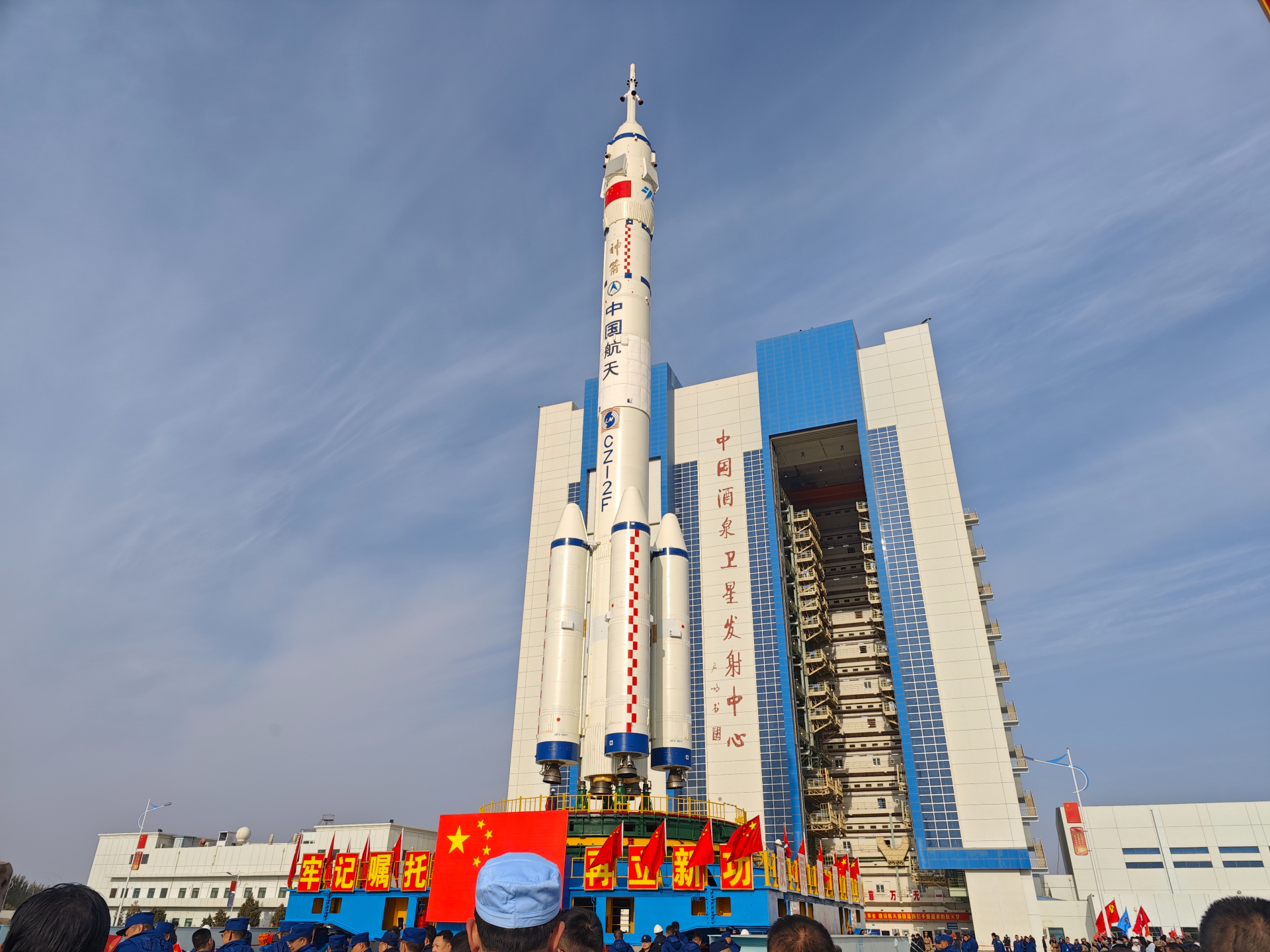 A large rocket stands next to a couple of tall buildings with the red Chinese flags in the front