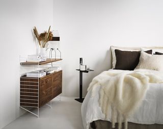 modular storage in a bedroom, storage with four drawers and shelving, attached to wall, black side table, bed with white bedding, sheepskin and brown pillows