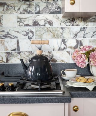 A kitchen countertop with a black granite surface, cream cabinets, and marble subway tiles, with a black kettle on the stovetop and cookies and roses on the counter