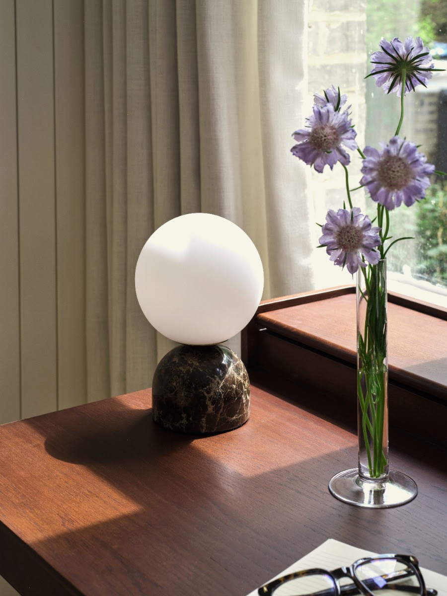 Image of a globe table lamp with a marble base sitting on a warm wooden desk with sunlight streaming in, and a clear, skinny vase of purple flowers.