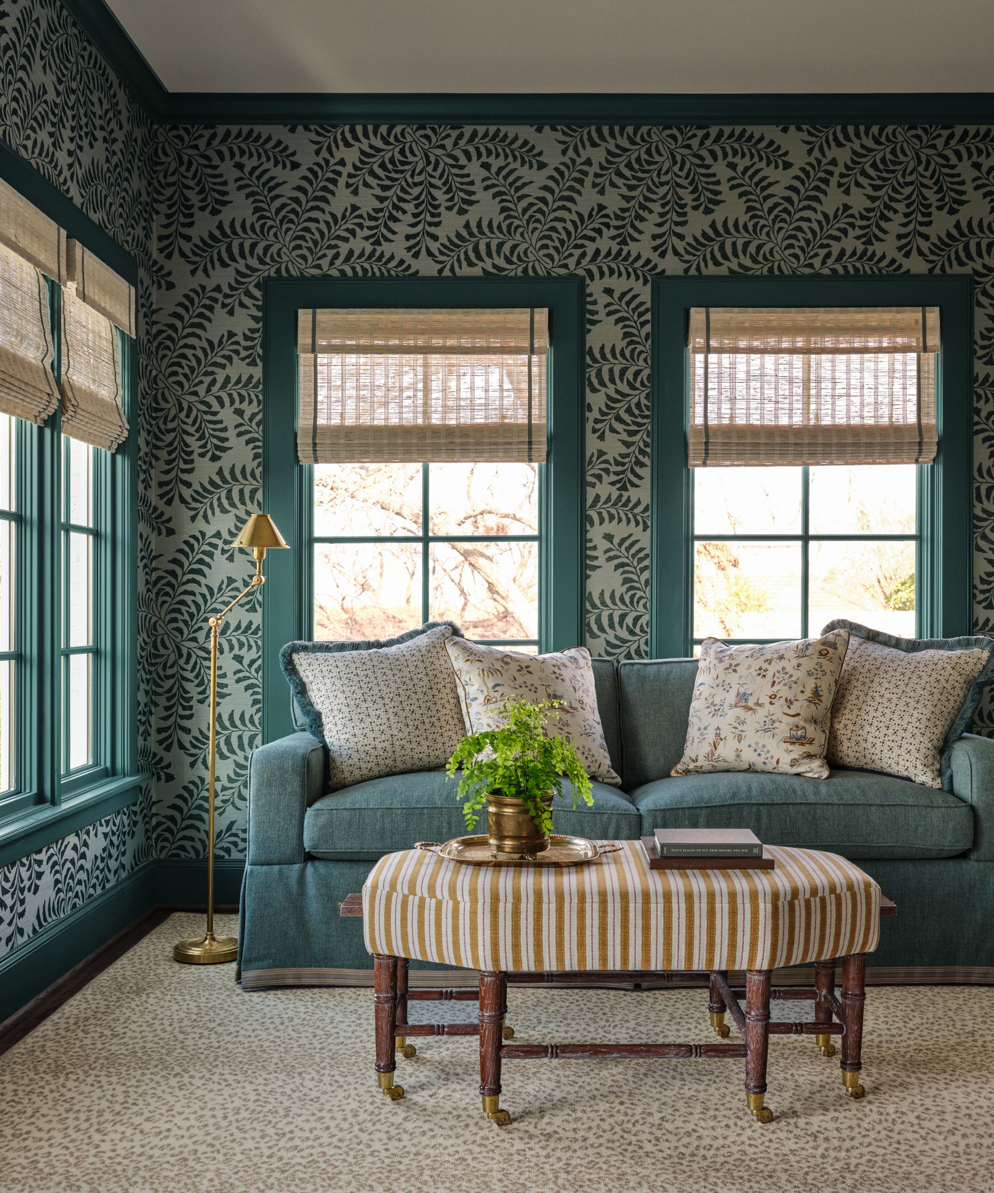 Living room with botanical blue wallpaper, blue window treatment, blue sofa and floral cushions, striped ottoman, and gold floor lamp