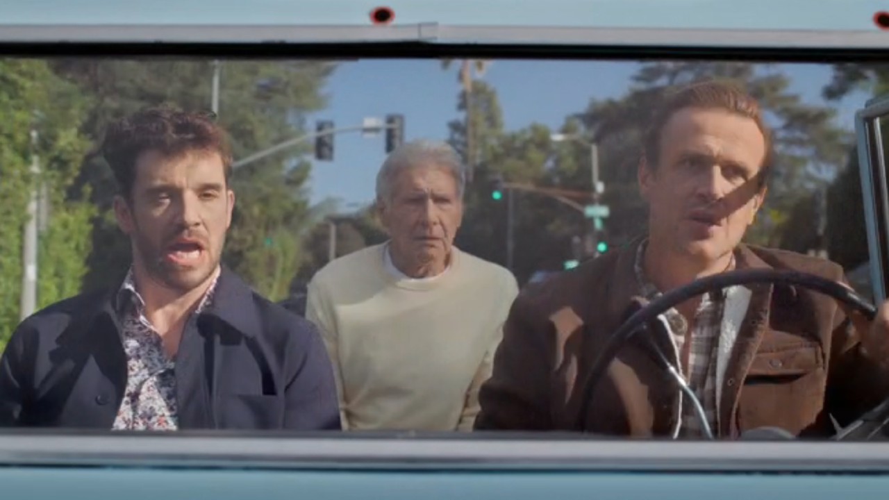 From left to right: Michael Urie sitting in the passenger seat singing, Harrison Ford sitting in the back looking confused and Jason Segel driving and singing. 