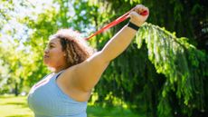 woman standing sideways wearing blue vest holding a resistance band overhead in a sunny park