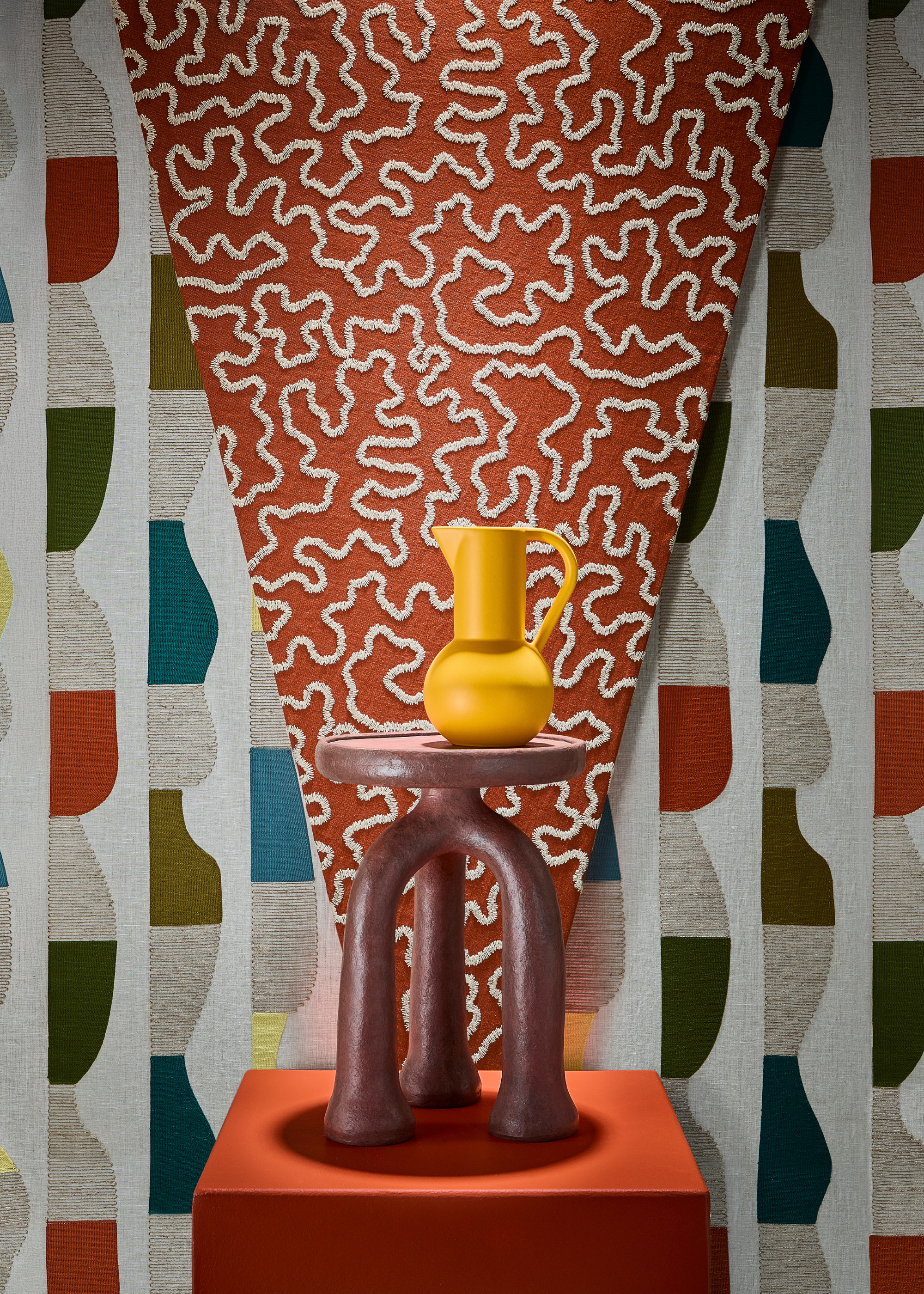 a design showcase featuring a modern side table with three clay-like legs in a browny-red colour on top of a red pedestal, with a vibrant yellow jug sitting on top of the table and against a backdrop of two contrasting patterned wallpapers