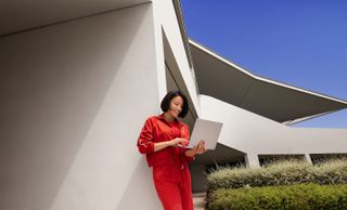 A woman in red clothes with black hair is holding a Snapdragon-powered laptop outside a white building near some shrubbery. It looks like a nice day with a deep blue sky