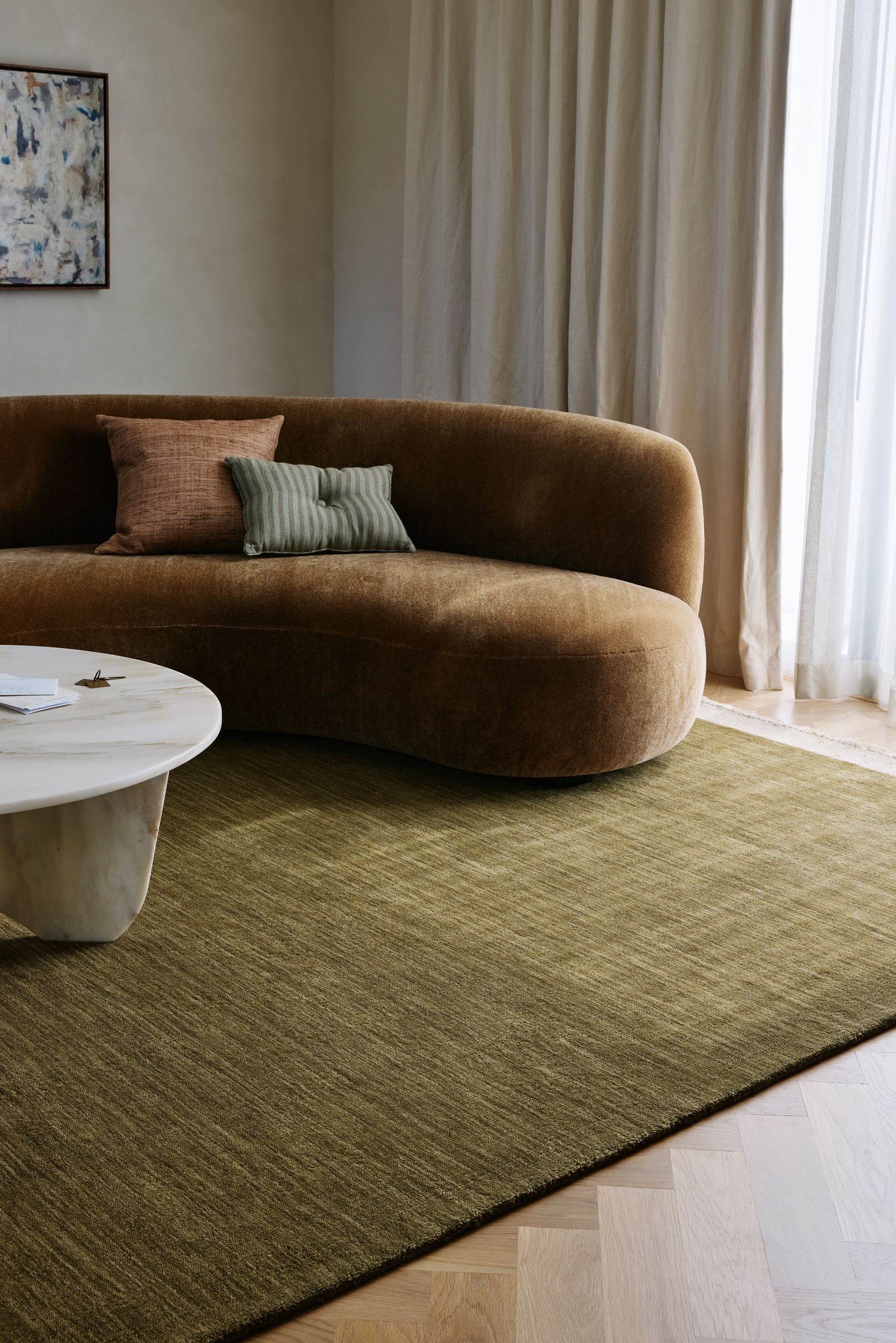 Image of a living room with an olive green rug, a curved light brown sofa and a white stone round coffee table.