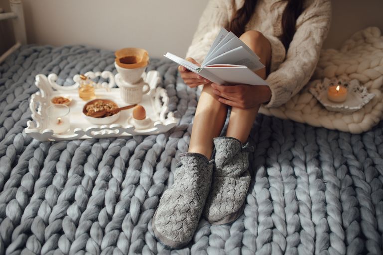 Woman with long hair drinking hot coffee and reading book in bed. Woman in woolen socks and sweater sitting on wool chunky merino plaid.