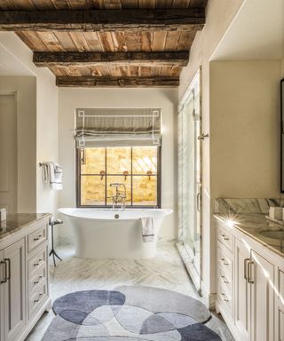 Large bathroom with rustic wooden ceiling, looking towards a bath beside a window, cream painted walls and fitted cabinets, marble flooring, rounded, abstract rug in blue and gray on floor
