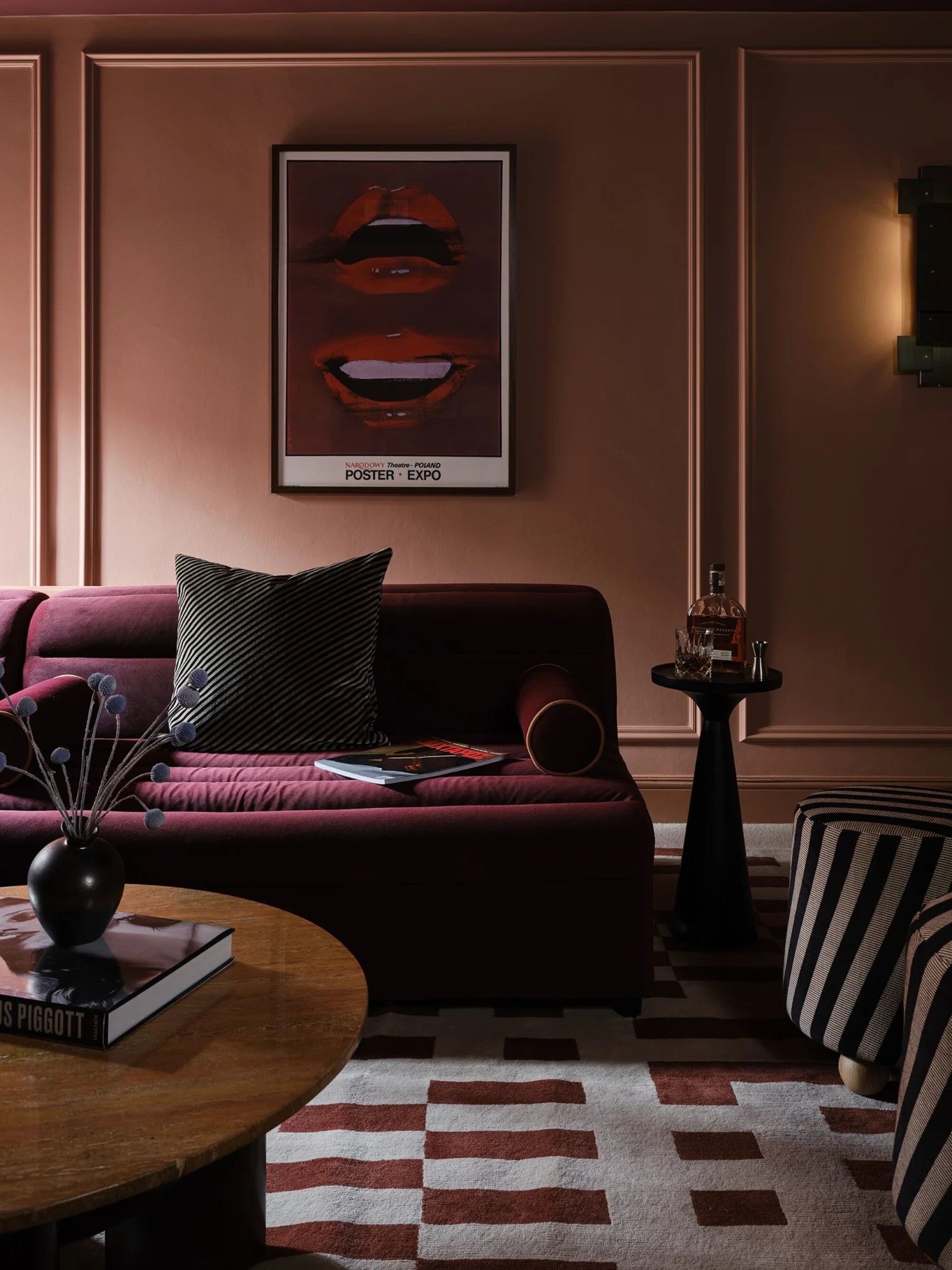 Image of a purple sofa in a dark living room where the walls are painted in a dark, dusty pink. There is a small drinks table beside the sofa and two black and white striped stools.