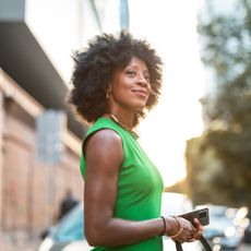 best serums for mature skin - black woman with Afro hair smiling softly, wearing sleeveless green slim-fitting dress and red lipstick and gold jewellery, holding her phone 