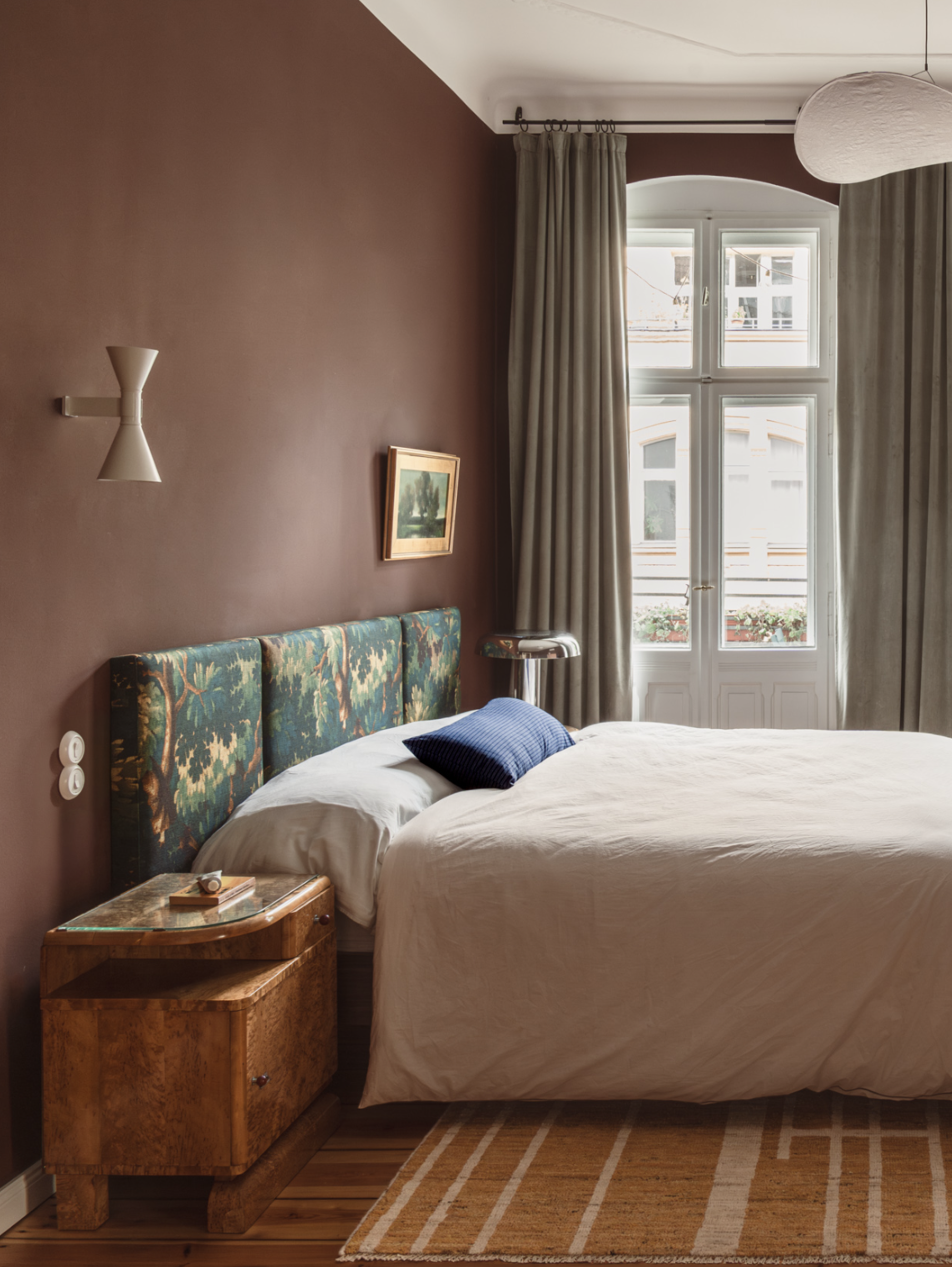 bedroom with brown walls, white ceiling, doors opening to balcony with tall curtains, upholstered tapestry headboard, off-centre artwork, white wall sconce over timber bedside table, and striped jute rug on timber floorboards