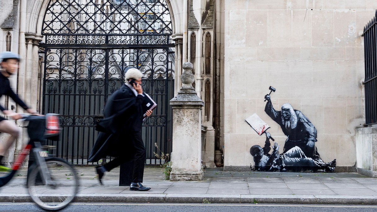 A photograph captures a person in a judge&#039;s robe walking while on a phone call, passing by a Banksy mural on a building wall that depicts an old, robed man wielding a hammer over a child holding a book.