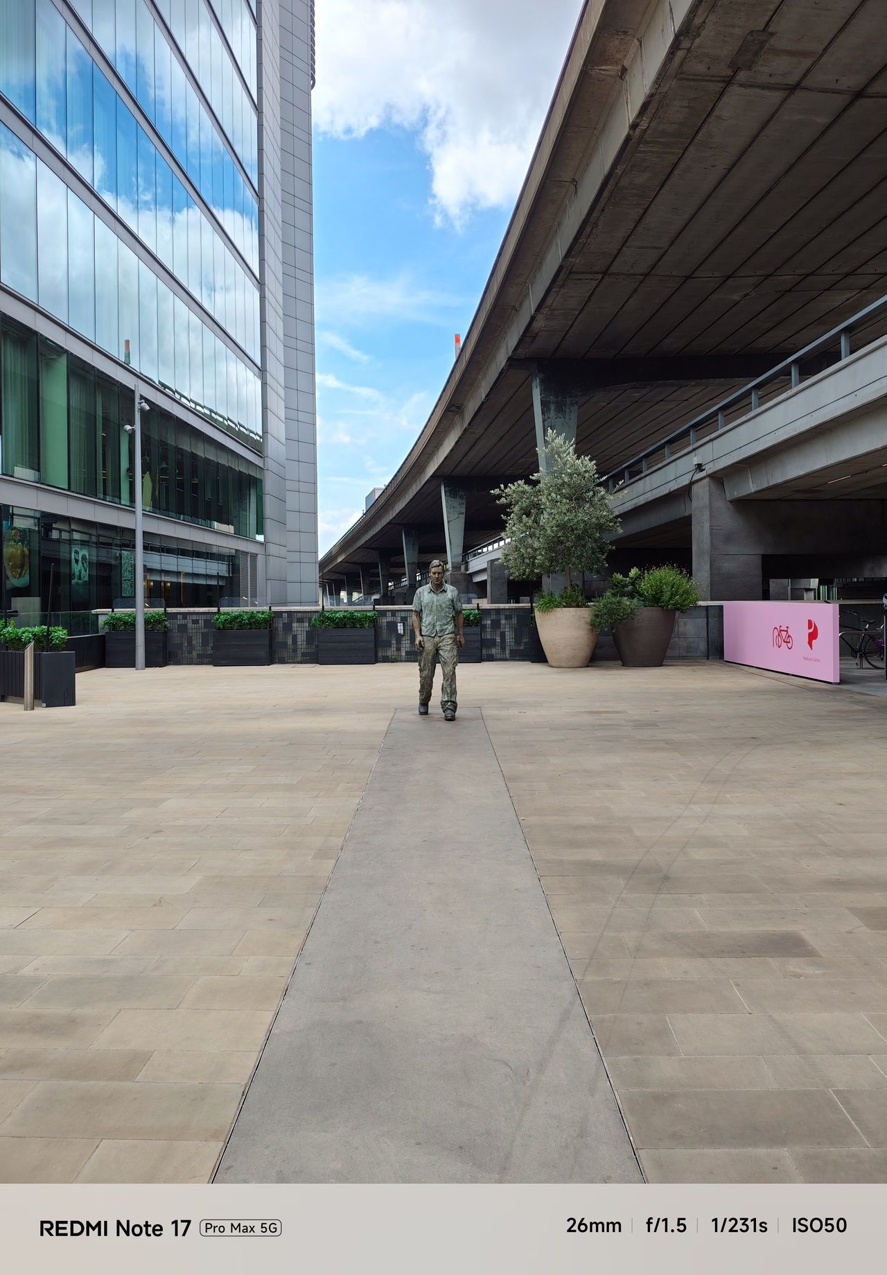 Urban plaza with a walking man sculpture centered between a modern office building and elevated roadway.
