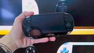 Hand holding PS Vita with Anbernic RG Vita handheld teaser on screen in backdrop.