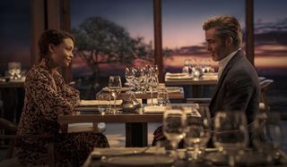 All The Old Knives - Celia Harrison (Thandiwe Newton) and Henry Pelham (Chris Pine) sit across from each other at a table for two in a fancy restaurant. She is wearing a black dress with a coral-coloured pattern, he is in a suit with an open-necked white shirt. There are several empty wine glasses on the table in front of them, and the view out of the window behind them is of a pink-hued sunset. They are smiling at each other.
