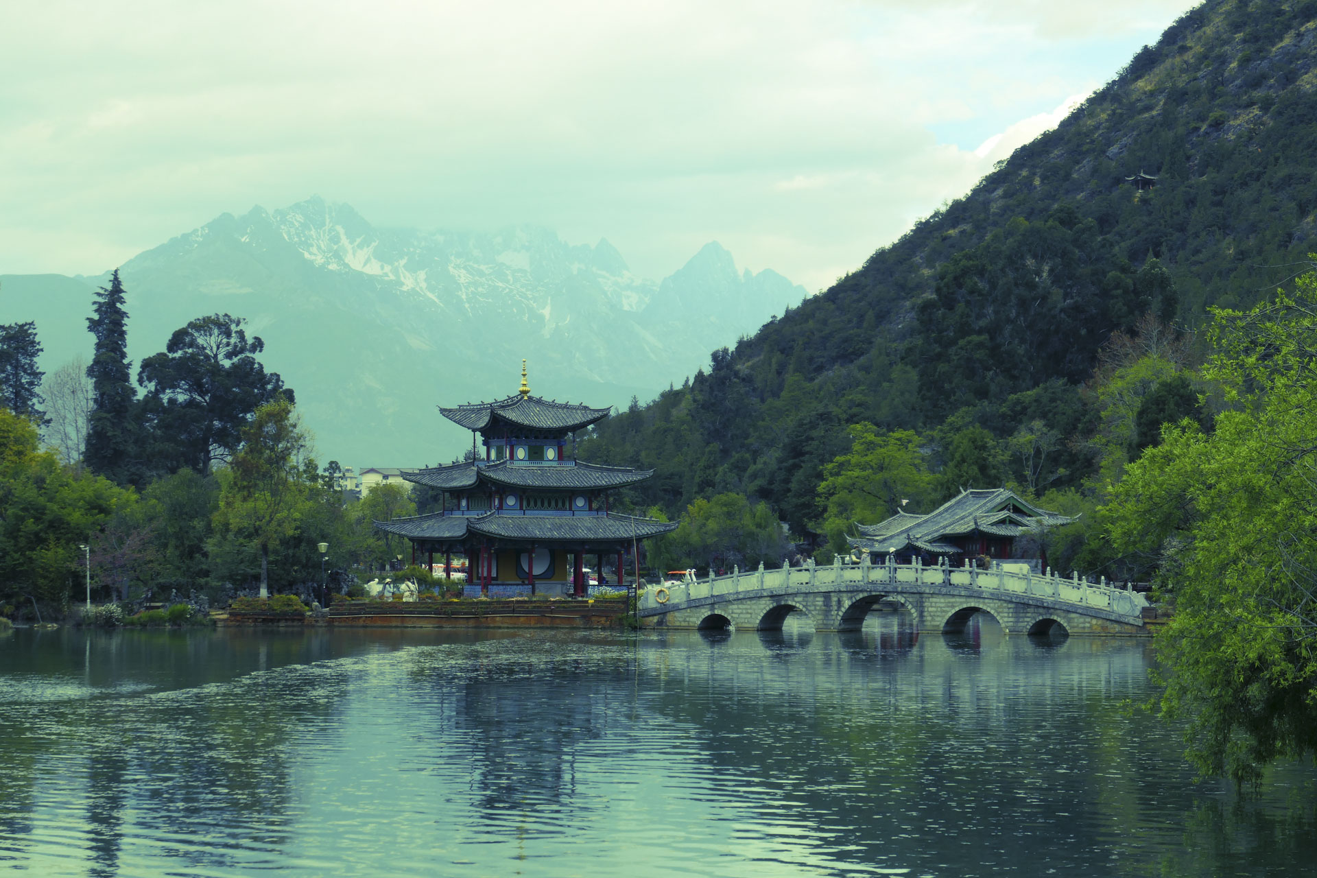 A Chinese temple by the side of a lake with an arched bridge leading up to it and both are reflected in the still water, in front of a snow covered mountain. A creative filter is applied to the image