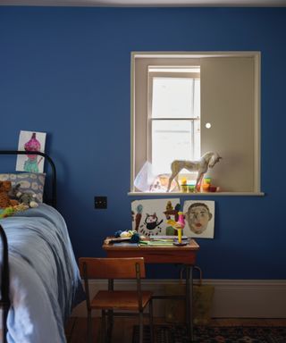 child's bedroom with dark blue wall, window, small desk with artwork on wall, corner of bed with light blue bedding