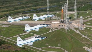 four white jets fly above a giant orange rocket on a launch pad below