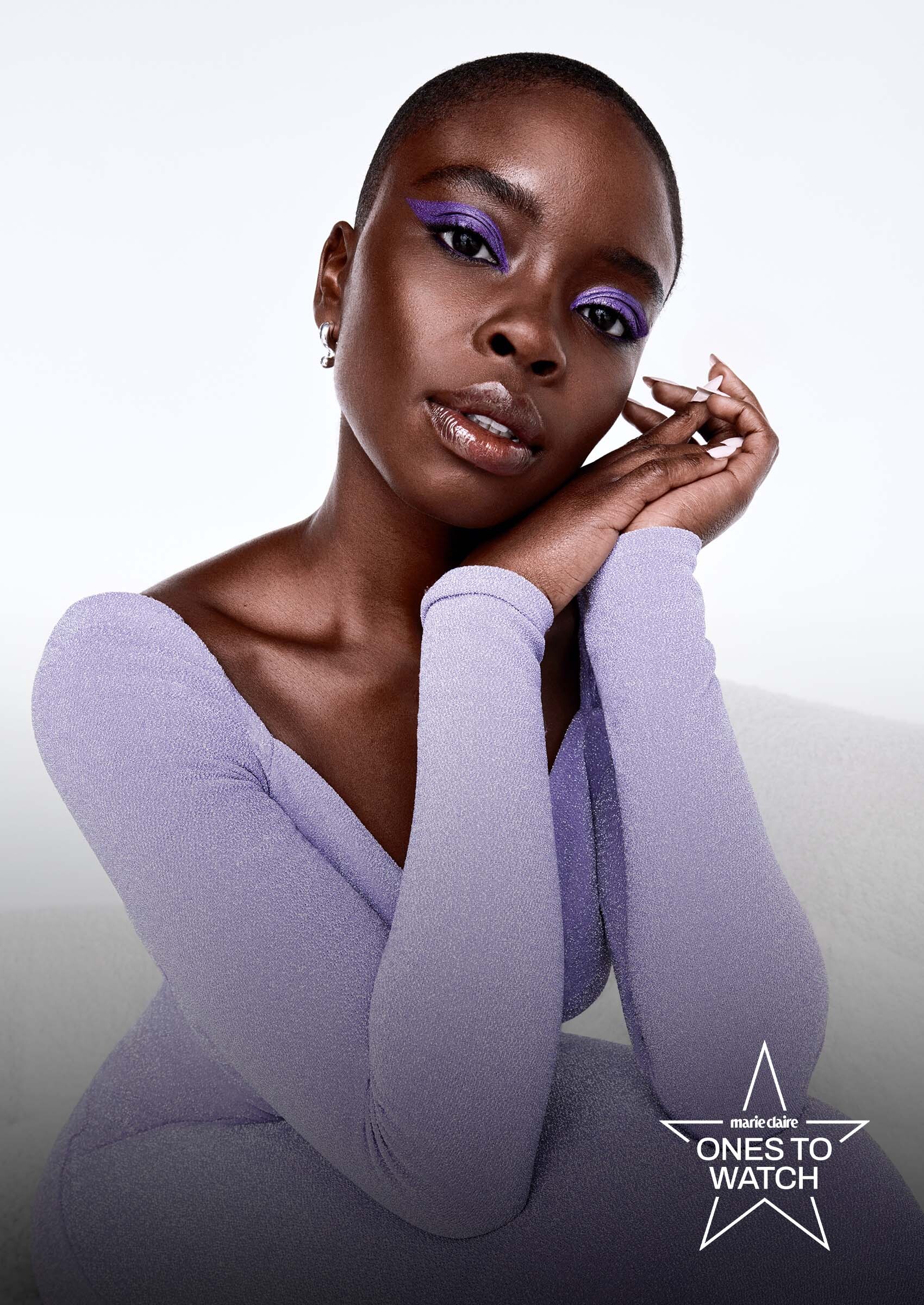 actress joy sunday poses resting her face on her hands and wearing a purple dress with purple eye shadow