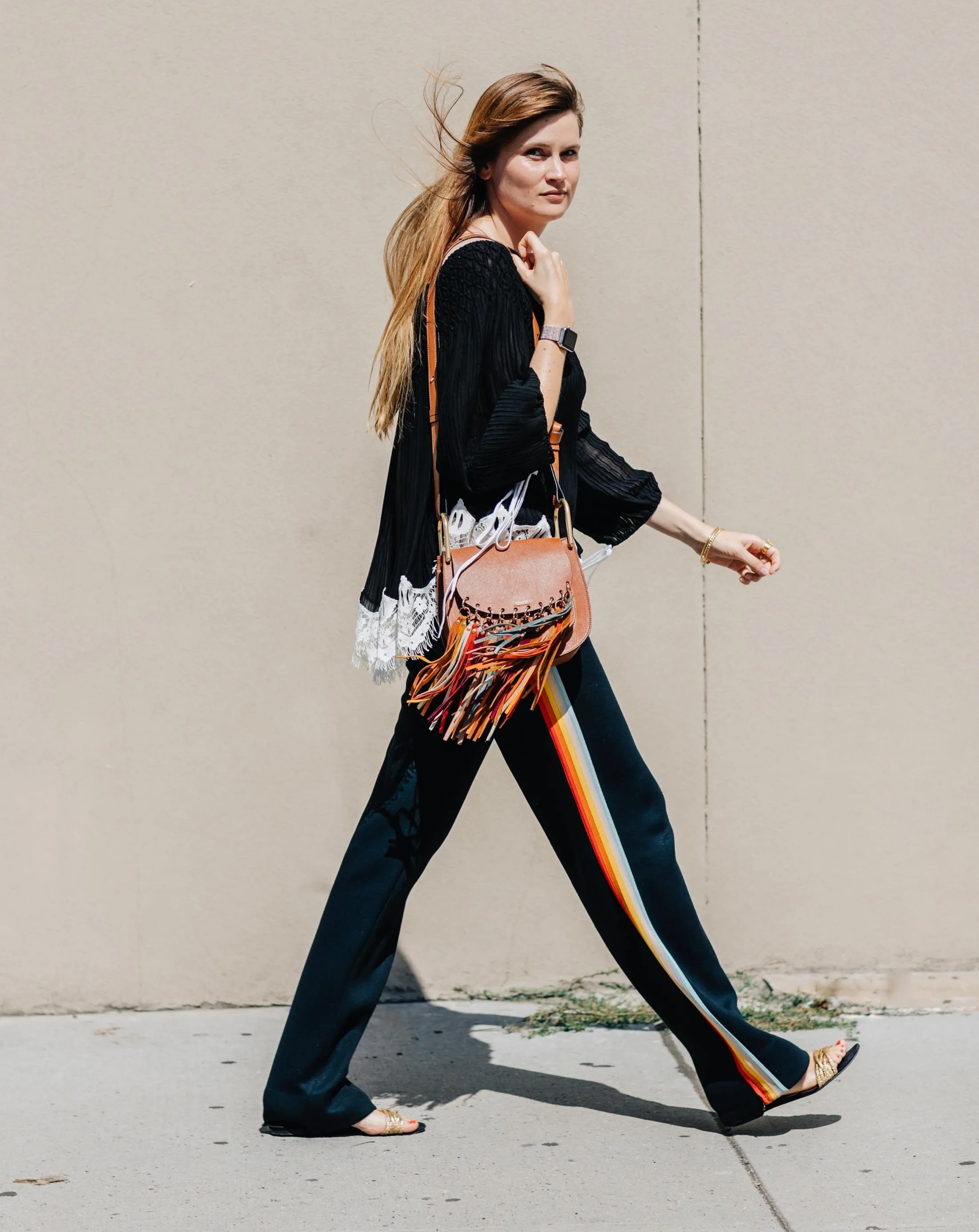 Street style shot of Eugenie Trochu wearing a lace-hem top, tan Chloe bag with rainbow fringe, rainbow stripe track pants, and sandals
