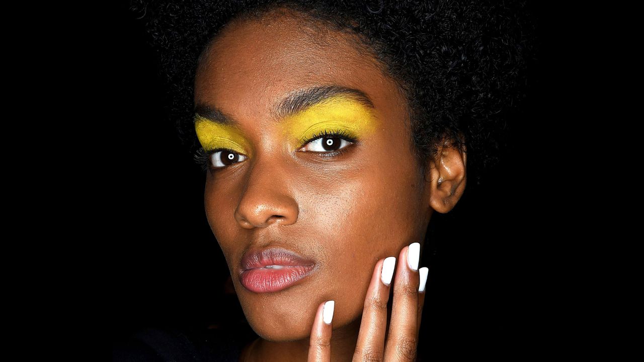 A black woman holds her fingers to her face, showcasing her white nail polish.