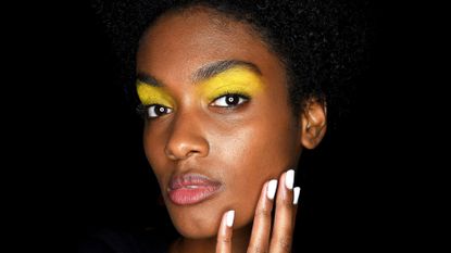 A black woman holds her fingers to her face, showcasing her white nail polish.
