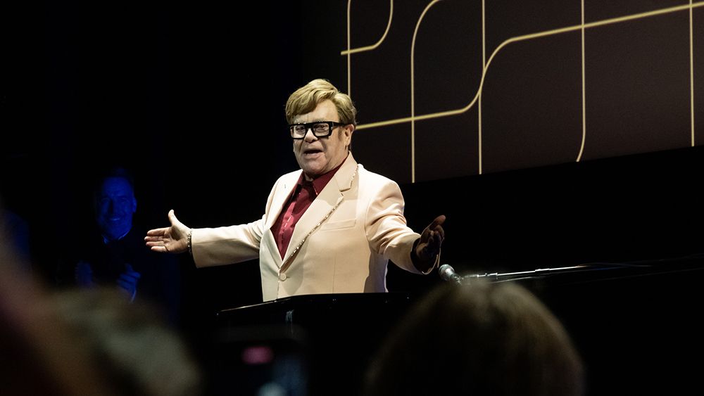 Elton John in a cream blazer with his arms out 