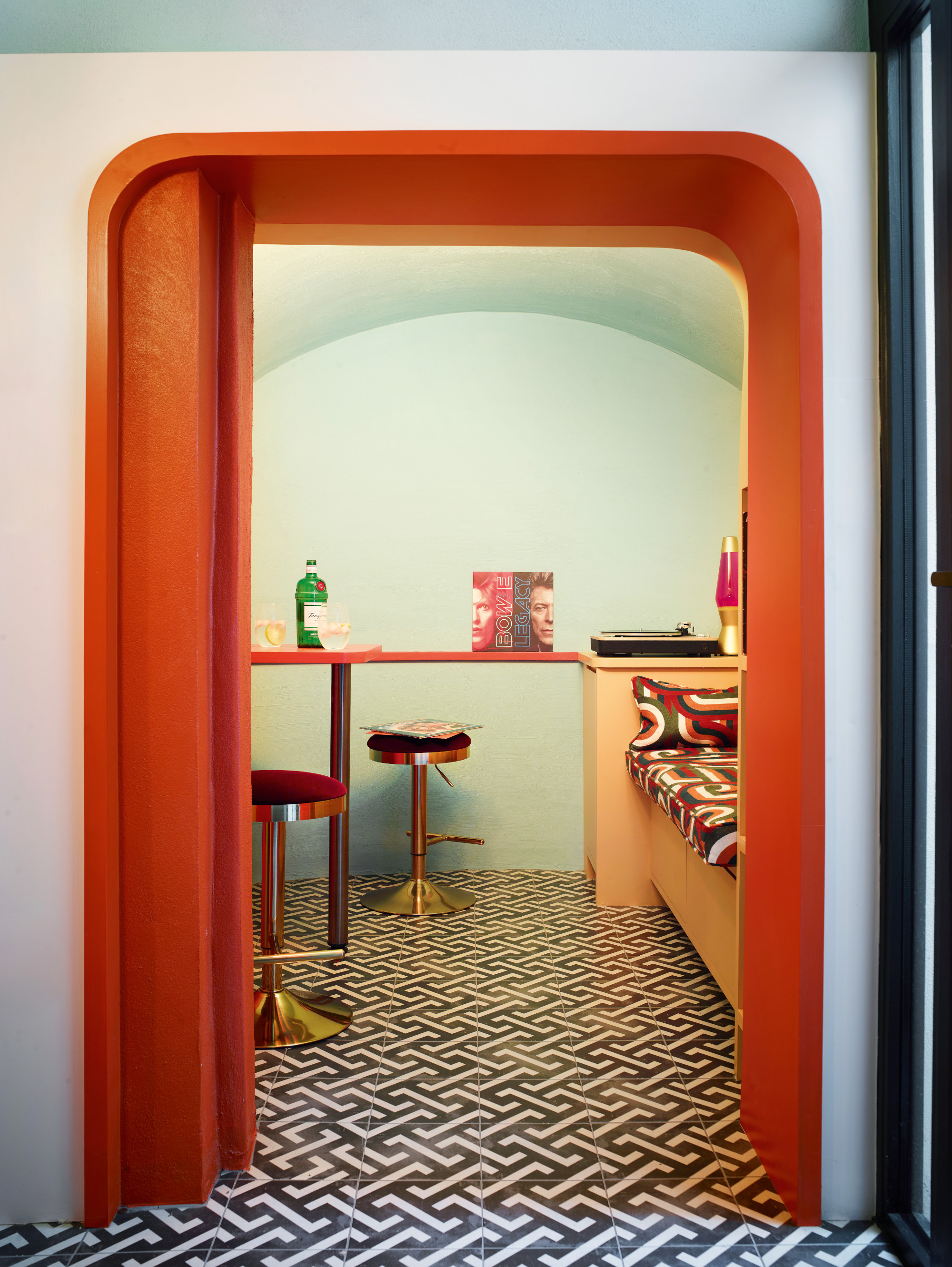 View into music room with green-blue walls, black and white geometric tiled floor, red and chrome bar stools around a slim red bar table, and bench with psychedelic green, red and white pattern