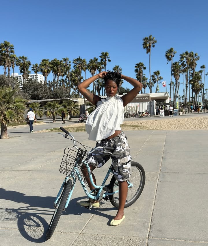 A woman on a bike wearing baggy camo shorts, a white one-shoulder blouse, and yellow Tabi flats.